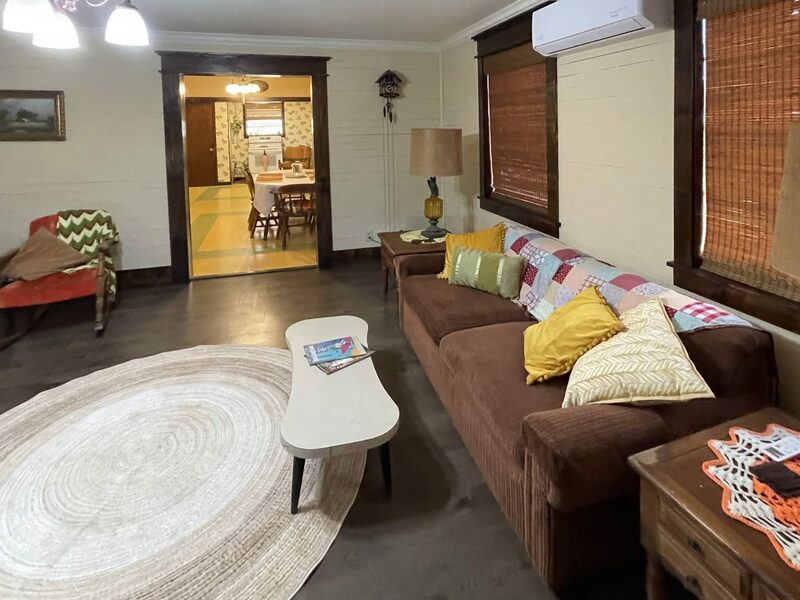 A living room with dark plank flooring: a brown sofa with yellow and green throw pillows, a small white coffee table and a red armchair with a green blanket, grouped around a large oval woven rug. Two windows with woven blinds and a wall-mounted air conditioner are on the right, and a doorway at the back opens to the dining area.